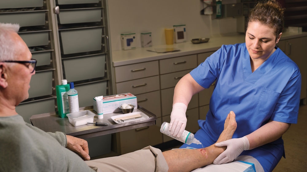 healthcare professional treating a wound on an elderly patient's lower leg in a clinical setting. The professional is applying Prontosan’s cleansing solution