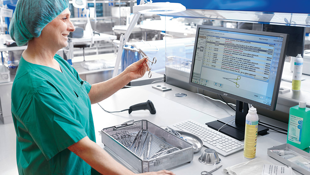 A female healthcare professional in scrubs stands by a computer, holding a surgical instrument and comparing it with an image on the screen